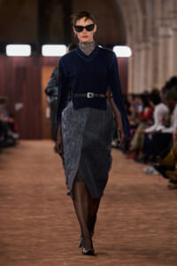 Model walking a runway in a navy velvet blouse with a black belt over a gray lace wrap skirt, dark sunglasses and sheer sleeves.