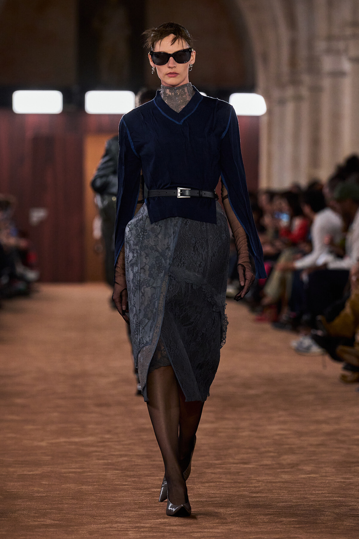 Model walking a runway in a navy velvet blouse with a black belt over a gray lace wrap skirt, dark sunglasses and sheer sleeves.