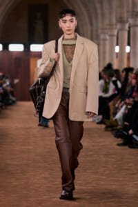 Model walking a fashion runway wearing a beige oversized blazer over a green knit sweater and brown leather pants, carrying a black leather bag.