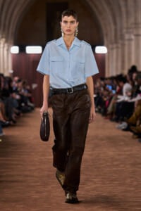 Runway model wearing a light blue short-sleeve shirt with brown leather pants, a black belt, metallic boots, and a small dark clutch on the runway.