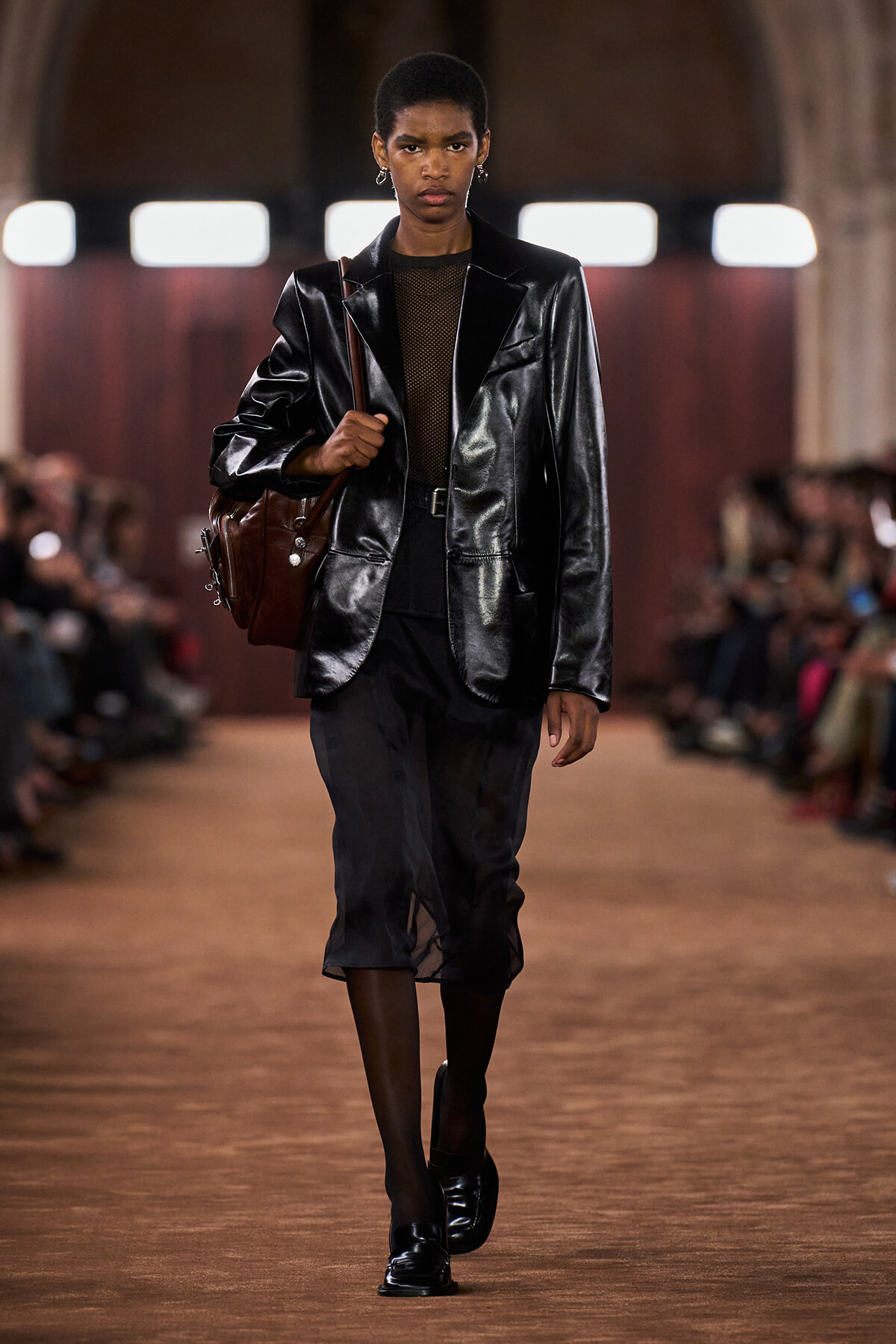 Model on a runway wearing a glossy black leather blazer, sheer top, black skirt, and a brown shoulder bagAssessing a fashion show look.