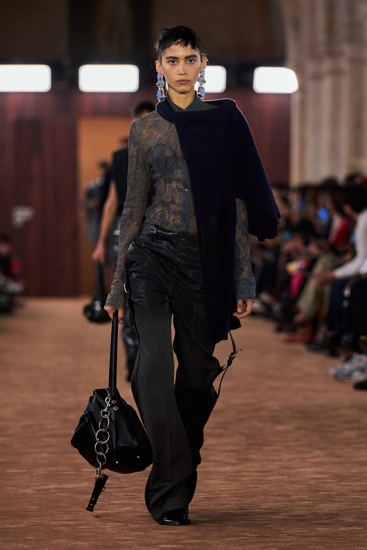 Model walks a runway in a sheer lace top, dark trousers, and a draped navy scarf, carrying a black bag with a silver chain.