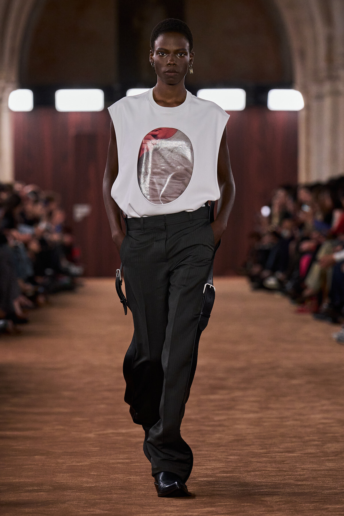 Model walking a runway in a white sleeveless top with a circular metallic print, paired with dark tailored pants.