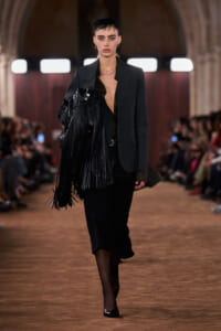 Model walking a runway in a dark blazer, velvet skirt, and a black fringe handbag.