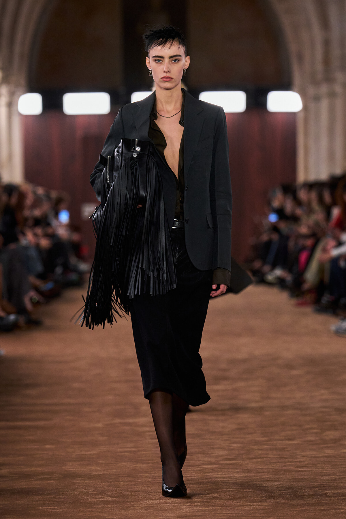 Model walking a runway in a dark blazer, velvet skirt, and a black fringe handbag.