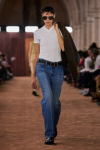 Model walking a runway in a casual white V-neck tee, blue jeans, black belt, sunglasses, with a brown leather jacket over the shoulder.