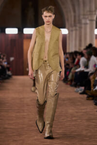 Model walks a runway in a sleeveless beige vest over a bare chest, metallic gold pants, and tall ornate beige boots; audience blurred in background.