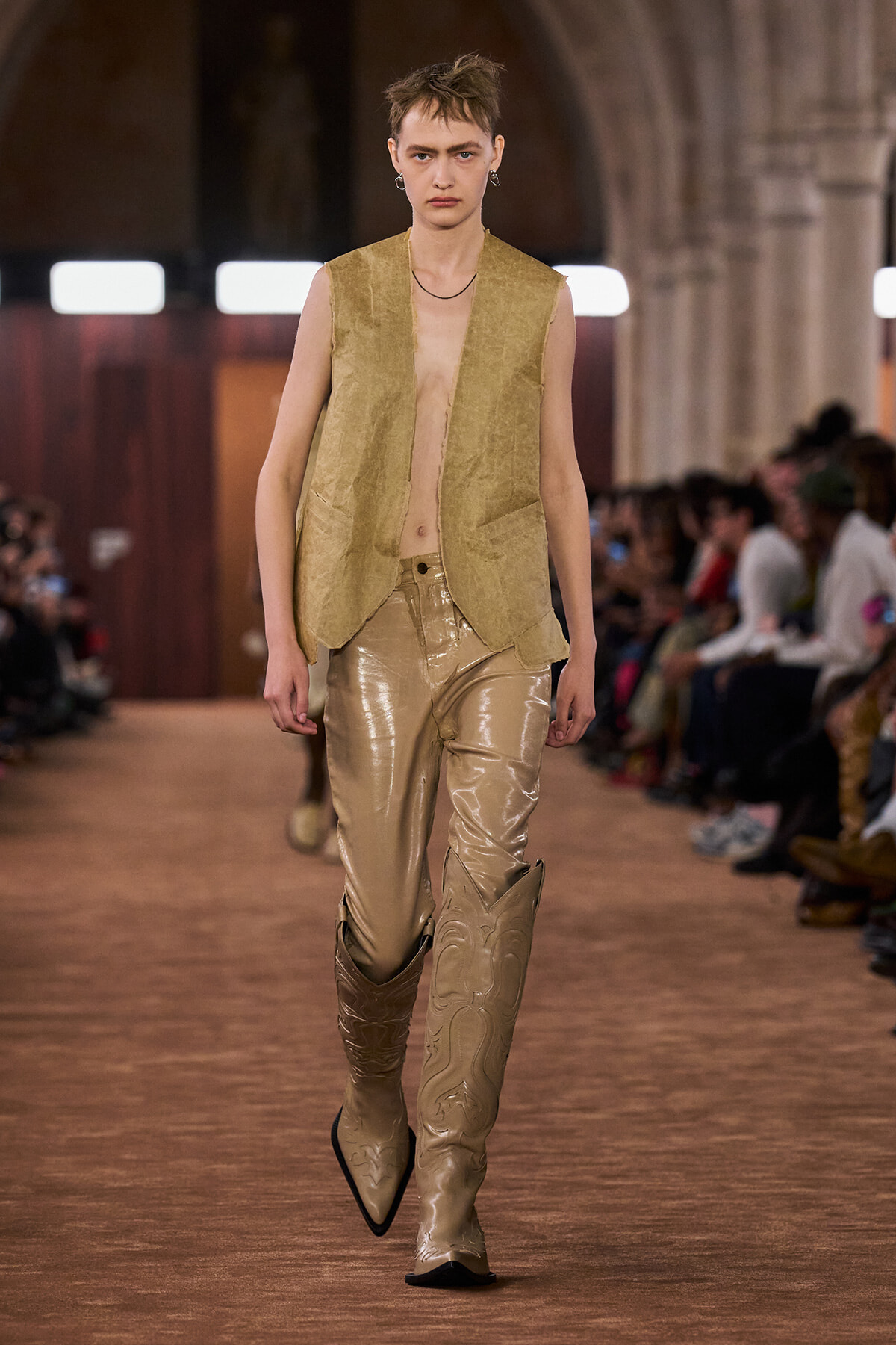 Model walks a runway in a sleeveless beige vest over a bare chest, metallic gold pants, and tall ornate beige boots; audience blurred in background.