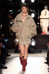 Model on a runway wearing a loose beige jacket over a textured mini dress, paired with red suede boots and a woven bag with tassels.