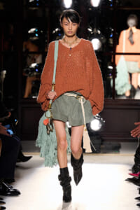 Model walking runway in a chunky rust knit sweater with olive shorts and a rope belt, carrying a mint fringe bag and wearing black slouch boots.