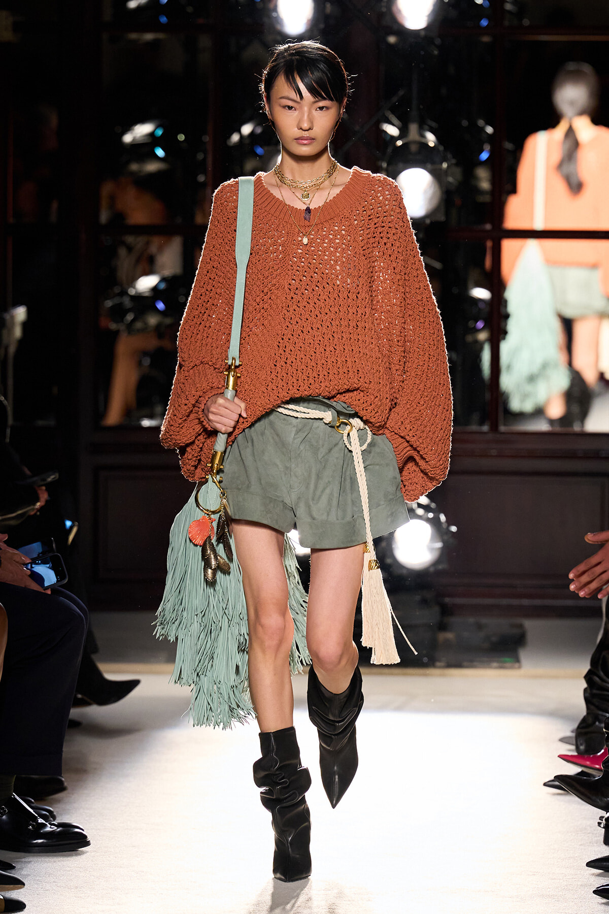 Model walking runway in a chunky rust knit sweater with olive shorts and a rope belt, carrying a mint fringe bag and wearing black slouch boots.