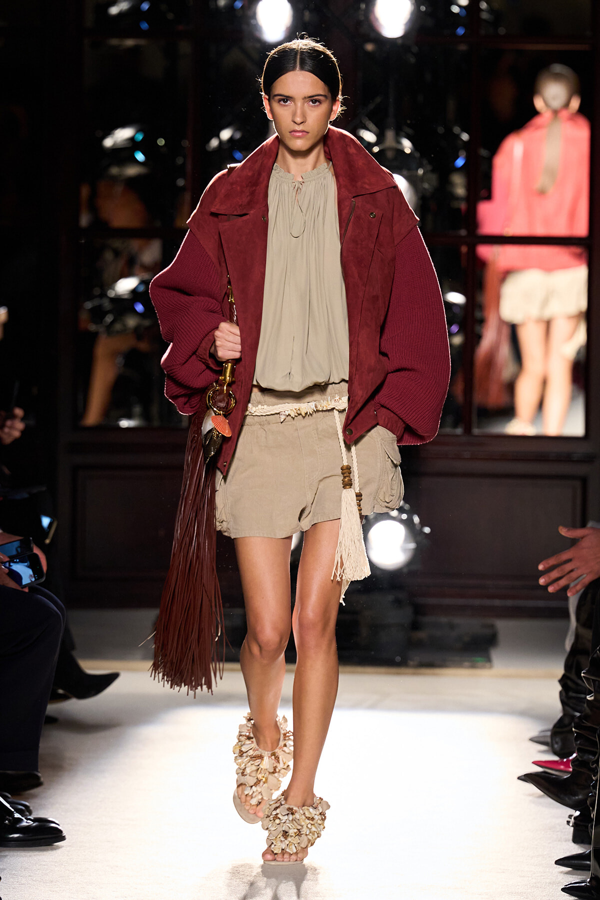 Model walks the runway in an oversized burgundy jacket over a beige blouse and tan shorts, carrying a fringed bag with long tassels and wearing floral-embellished sandals.