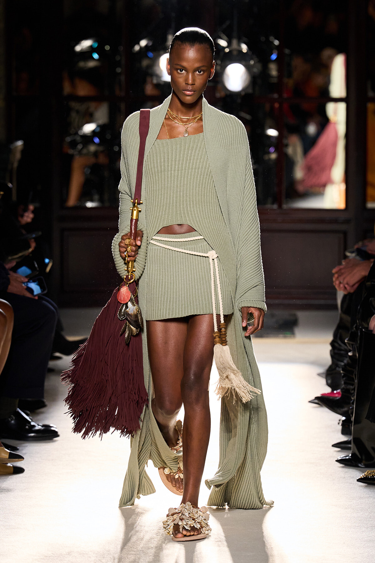 Model walks down the runway in a sage-green ribbed knit ensemble: a long cardigan over a cropped top and wrap-skirt with a white rope belt, paired with beaded sandals and a large burgundy fringed bag accessorized with tassels and jewelry.