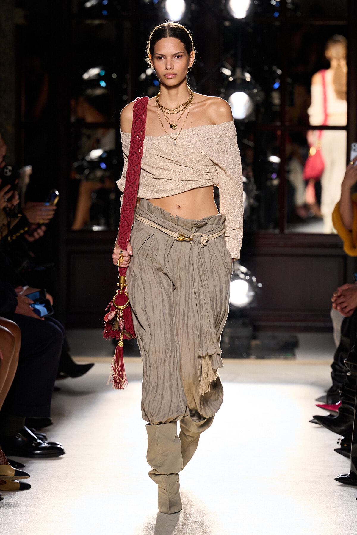 Model walking the runway in a beige off-shoulder knit top, loose crinkled gray pants with a rope belt, and beige boots, carrying a red braided bag over the shoulder.