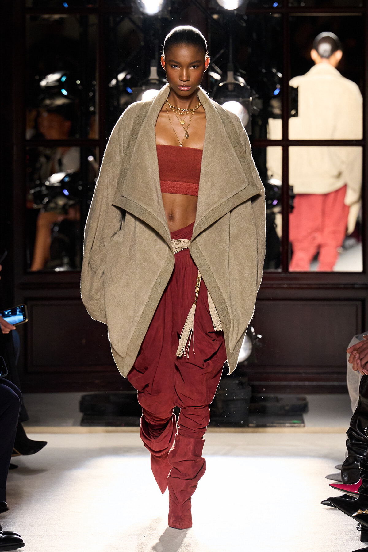 Model walking runway in a oversized beige cape over a red bandeau top and draped burgundy harem pants with tassel belt and matching boots.