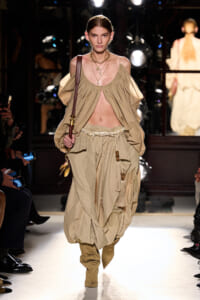 Model walking a runway in a beige draped two-piece ensemble with a cutout midriff and harem-style pants, accessorized with layered necklaces and a brown shoulder bag.
