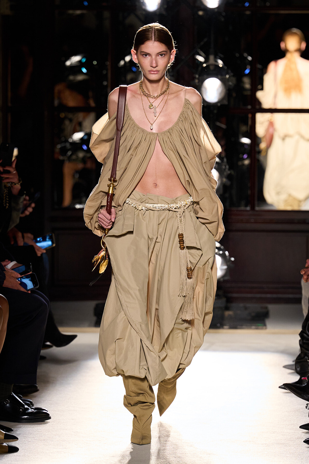 Model walking a runway in a beige draped two-piece ensemble with a cutout midriff and harem-style pants, accessorized with layered necklaces and a brown shoulder bag.