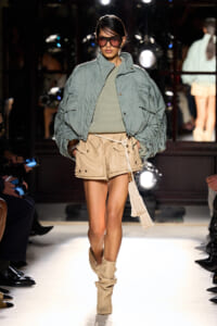 Model on a runway wearing an oversized light blue-green jacket over an olive knit top and beige cargo shorts with tan slouch boots, sunglasses on.