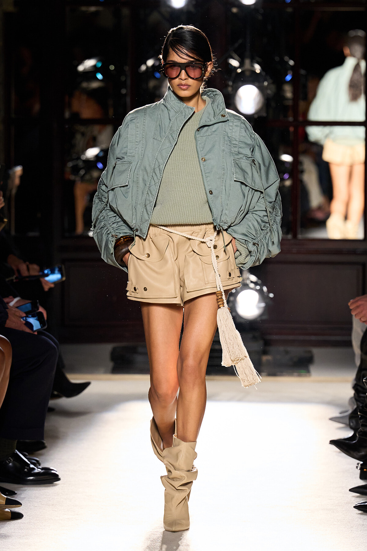 Model on a runway wearing an oversized light blue-green jacket over an olive knit top and beige cargo shorts with tan slouch boots, sunglasses on.
