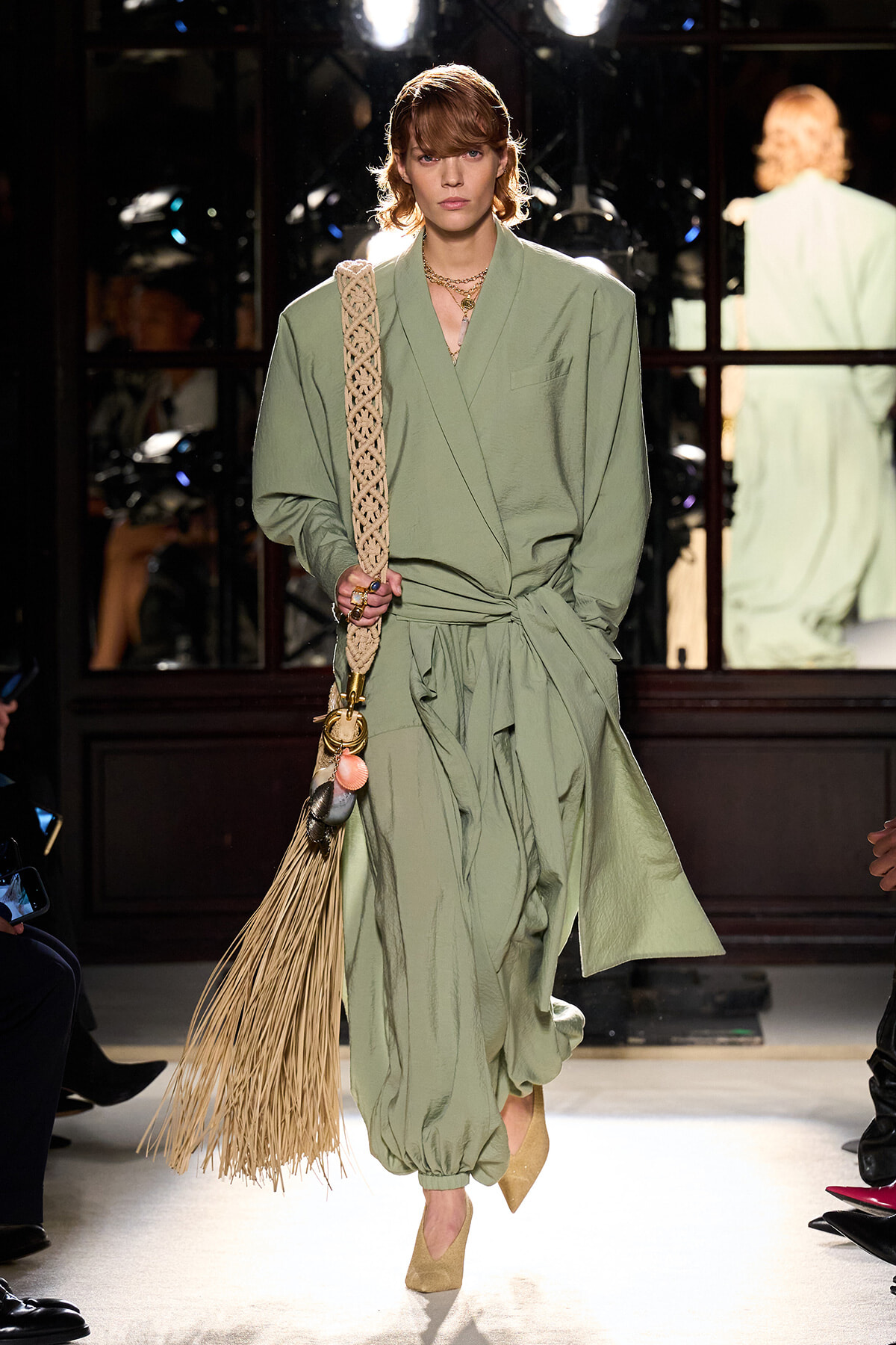 Model walking the runway in a sage-green, wrap-style jumpsuit with a braided fringe bag over her shoulder.