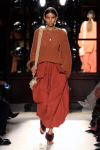 Model wearing a chunky orange-brown knit sweater and a voluminous rust skirt, walking a runway with a woven bag over her shoulder.