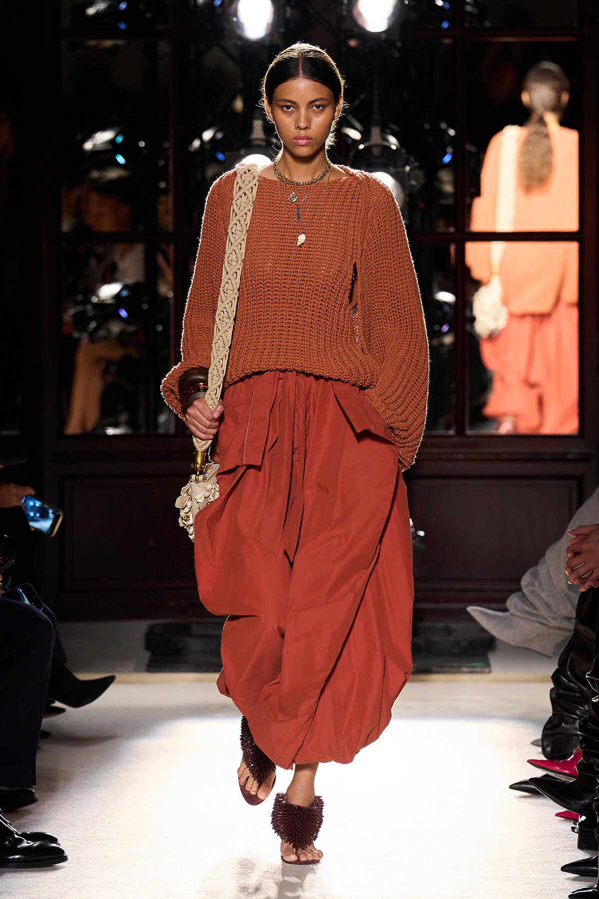Model wearing a chunky orange-brown knit sweater and a voluminous rust skirt, walking a runway with a woven bag over her shoulder.