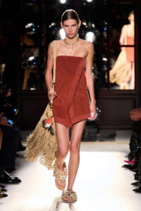 Model walks a runway in a rust strapless, wrap-style romper with a front panel; beige fringed bag slung over shoulder and layered gold necklaces. She wears floral-embellished sandals, and the scene is a fashion show with photographers and spectators in the background.