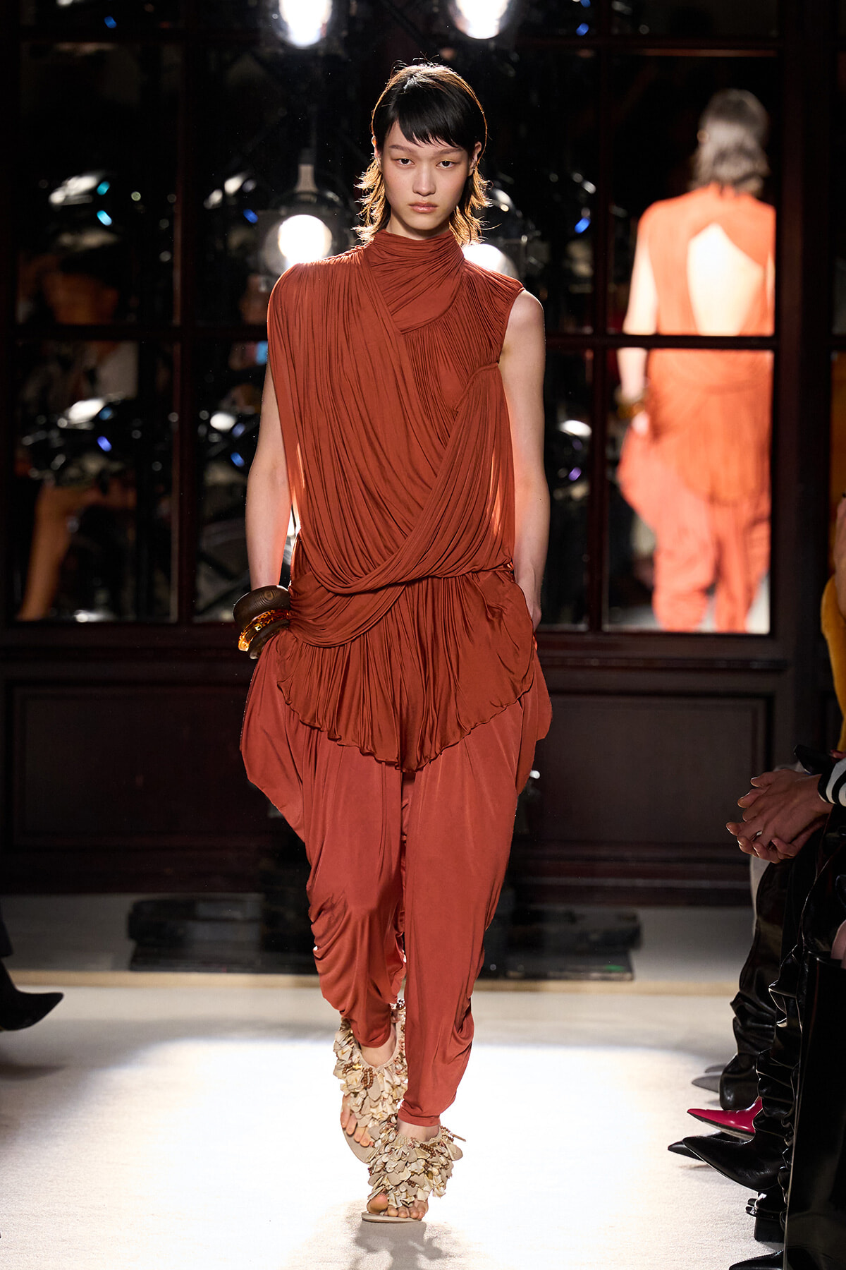 Female model walks a fashion runway in a rust-orange draped sleeveless top and matching harem pants with floral-embellished sandals, backstage mirrors visible.
