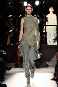 Model walking a runway in a sage-green, draped, ruched dress with matching slouchy boots and a small red fringed handbag.