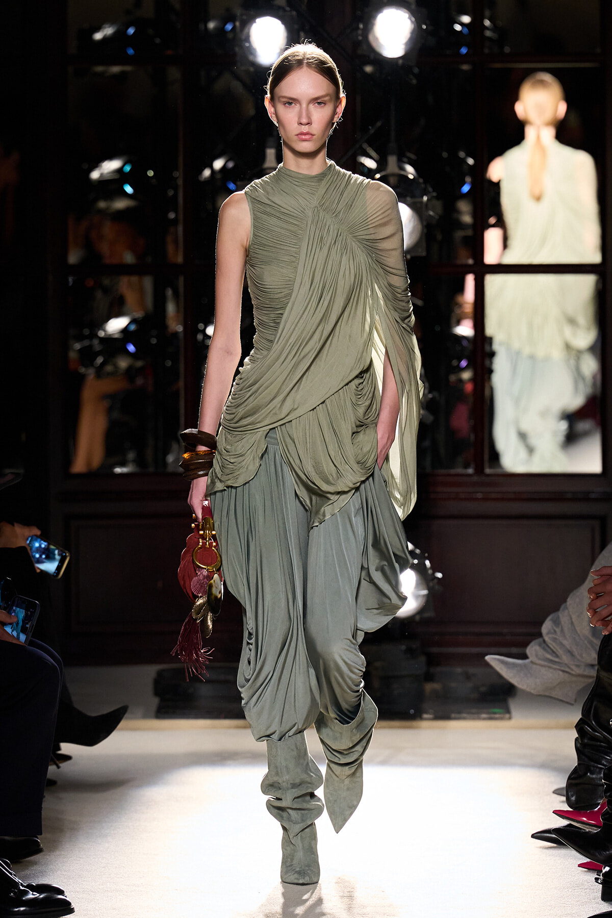 Model walking a runway in a sage-green, draped, ruched dress with matching slouchy boots and a small red fringed handbag.