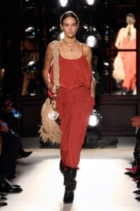Model walks runway in rust-red draped jumpsuit with knotted waist, accessorized with layered necklaces and a beige fringe bag over the shoulder; black knee-high boots visible.
