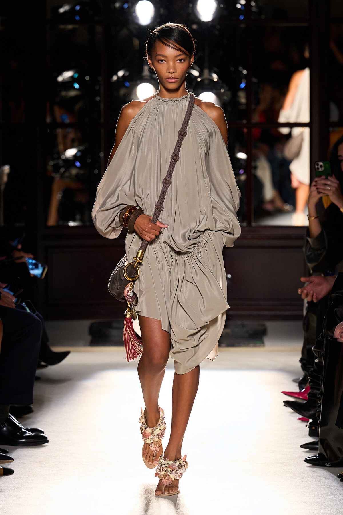 Model walking a fashion show in a taupe off‑the‑shoulder, voluminous dress with gathered waist, carrying a braided‑strap bag with tassels.