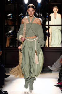 Model walking runway in sage-green, off‑shoulder knit set with wrapped halter top and voluminous pants, barefooted/in-beige fringe bag accessory
