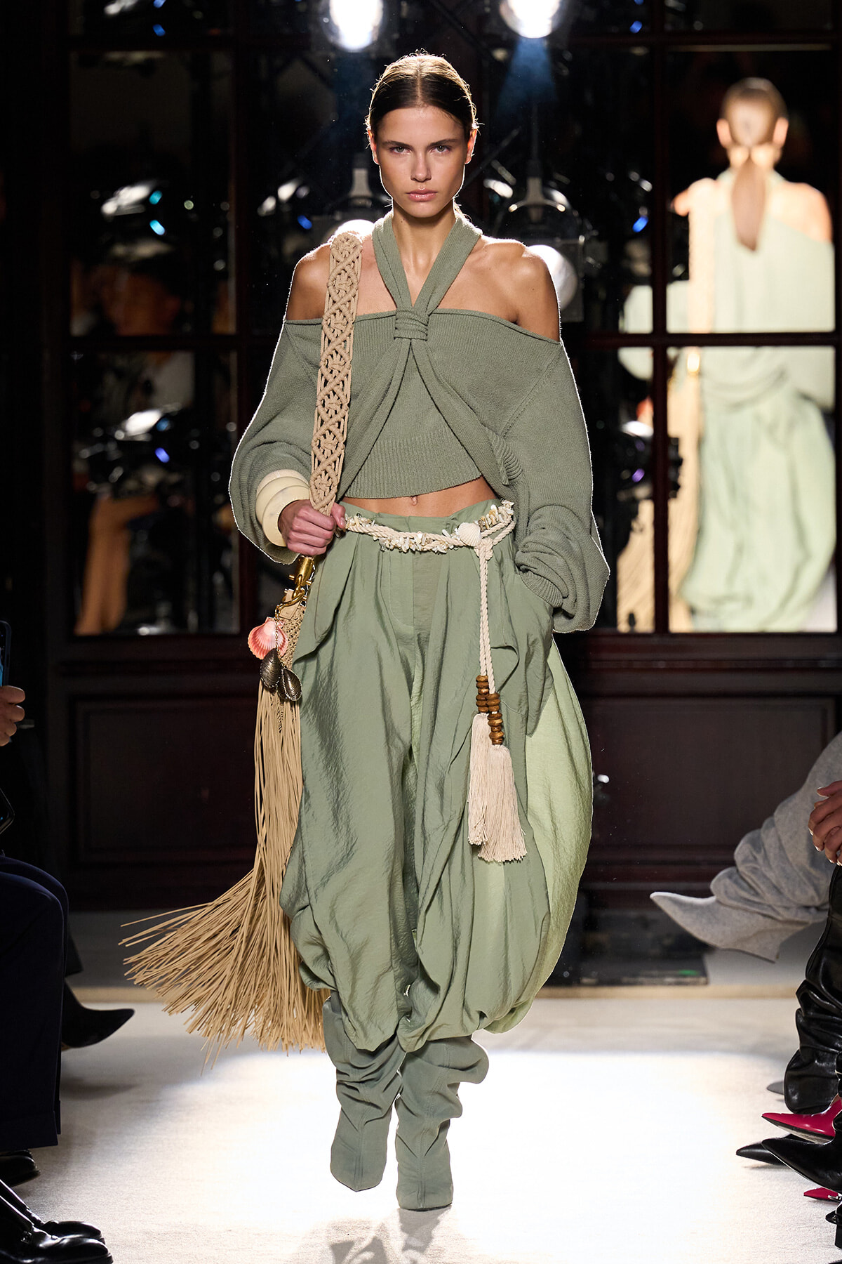 Model walking runway in sage-green, off‑shoulder knit set with wrapped halter top and voluminous pants, barefooted/in-beige fringe bag accessory