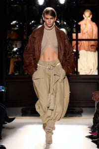 Model on runway in a cropped beige knit top, brown leather jacket, and baggy khaki cargo pants with a rope belt.