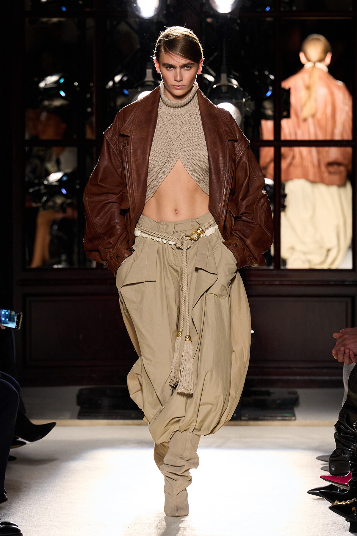 Model on runway in a cropped beige knit top, brown leather jacket, and baggy khaki cargo pants with a rope belt.