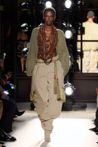Model walking a runway in a green knit sweater and oversized beige cargo pants, wearing multiple layered beaded necklaces.