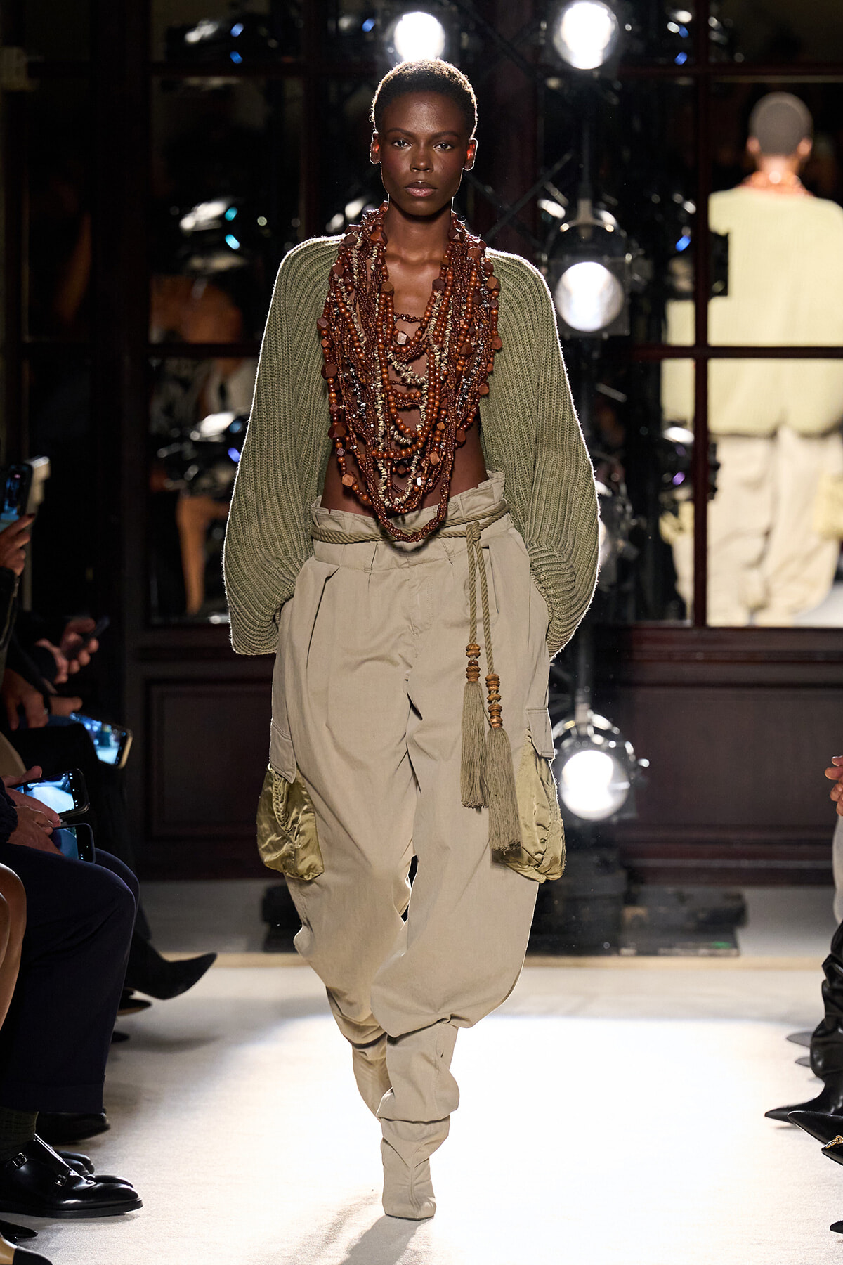 Model walking a runway in a green knit sweater and oversized beige cargo pants, wearing multiple layered beaded necklaces.