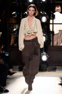 Model walks the runway in a cream oversized cardigan with a multi-strand beaded scarf, paired with brown draped trousers and tassel accents.