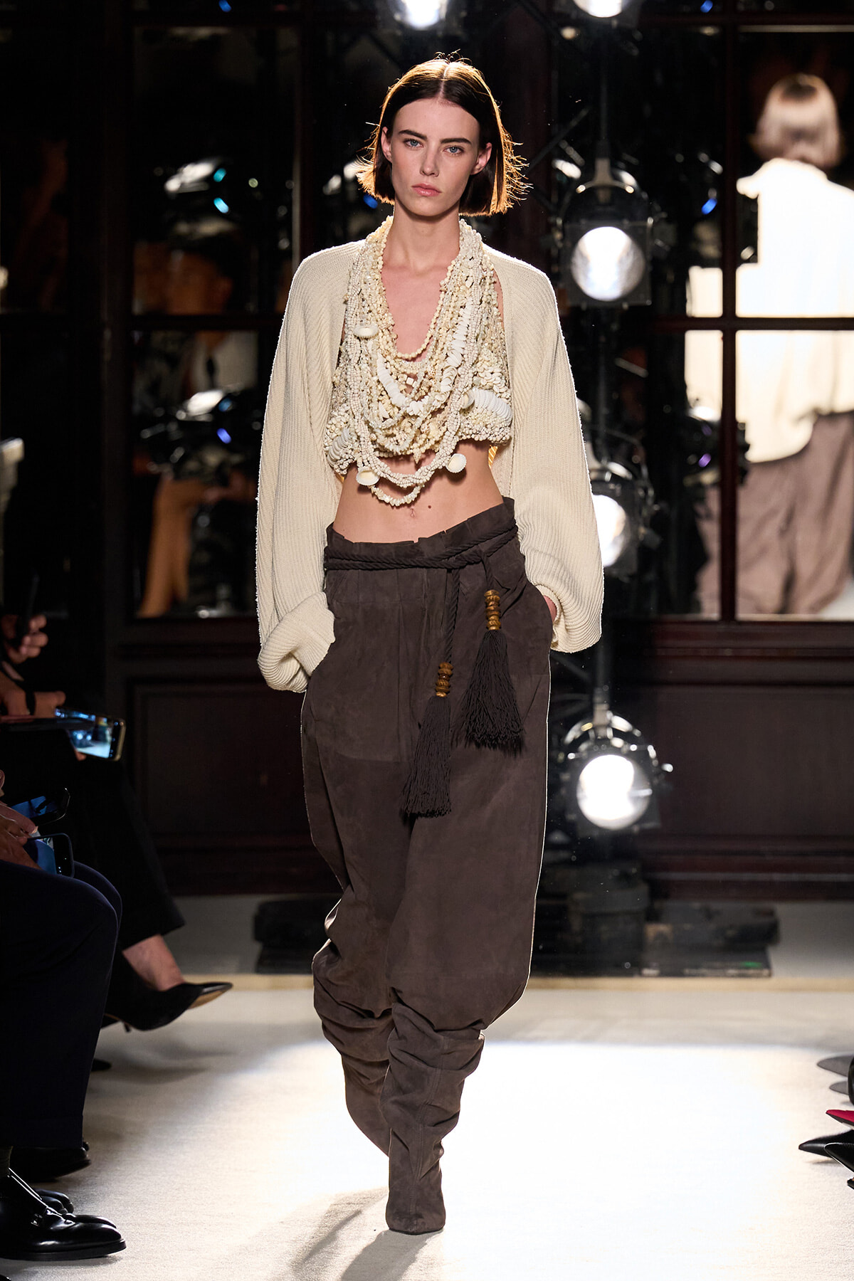 Model walks the runway in a cream oversized cardigan with a multi-strand beaded scarf, paired with brown draped trousers and tassel accents.