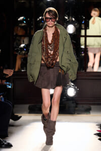 Model walking a runway in an oversized olive utility jacket, multiple brown beaded necklaces, brown shorts, and slouchy suede boots, with dark sunglasses.