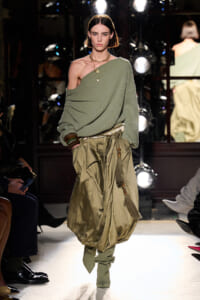 Model walks a fashion runway in a sage green one-shoulder knit top and oversized olive draped pants, with layered gold necklaces and bracelets.