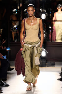 Model walking runway in a beige sleeveless top and olive green draped pants with a white woven belt and fringed bag.
