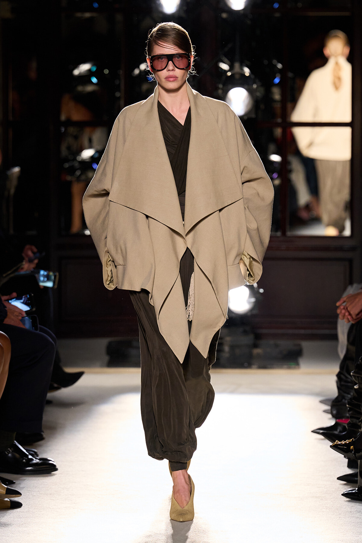 Model on a runway wearing a oversized beige cape coat over dark inner wear and olive pants with beige heels and large sunglasses.