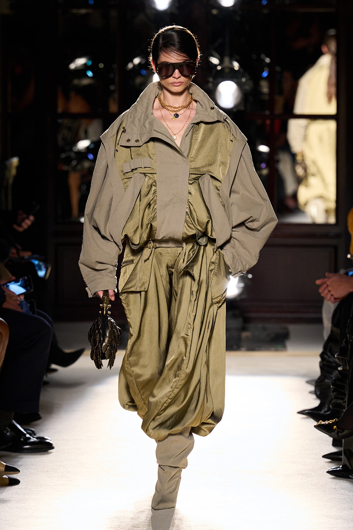Model walks a runway in an oversized khaki utility jacket with gathered waist, matching pants, large sunglasses, gold necklaces, and a small black textured handbag.
