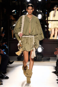 Model walking a runway in an olive-green, billowy dress with a rope belt and beige boots, carrying a woven bag with tassels and shells.