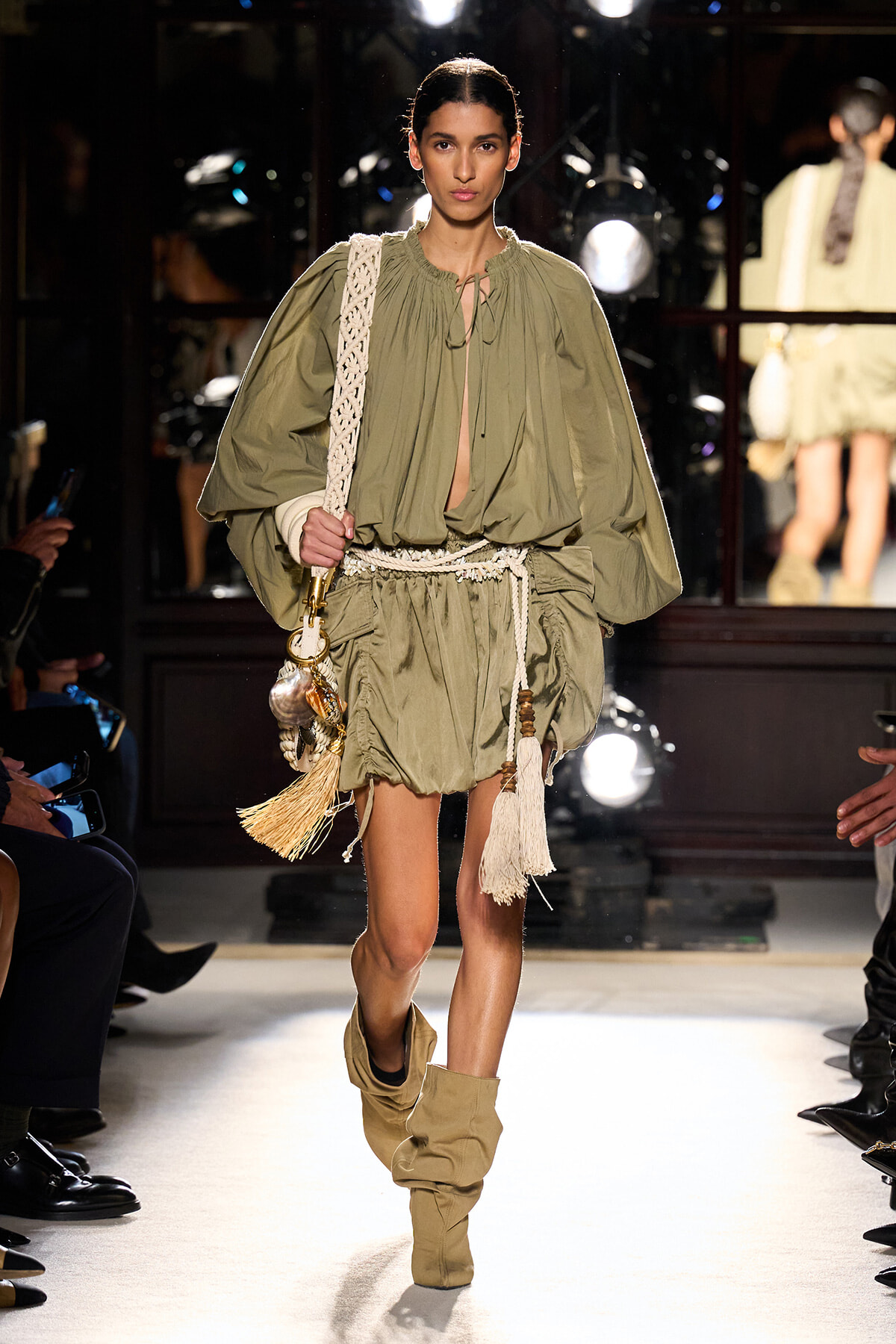Model walking a runway in an olive-green, billowy dress with a rope belt and beige boots, carrying a woven bag with tassels and shells.