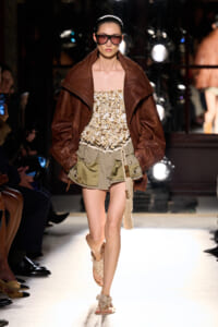 Model walks the runway in a large brown leather jacket over a beige beaded top and olive-green shorts, with dark sunglasses and sandals.