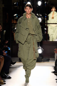 Model on a fashion runway wearing a loose olive-green utility jacket over a matching knit top and oversized cargo pants, accessorized with a rope belt and layered necklaces.