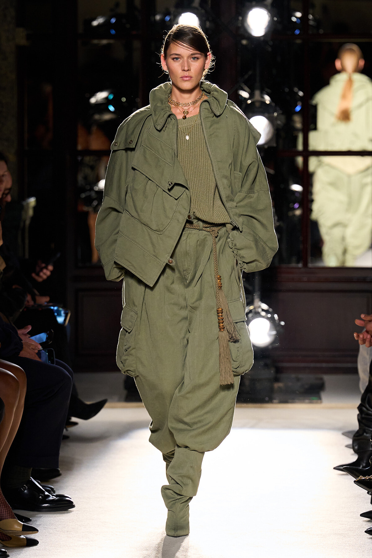 Model on a fashion runway wearing a loose olive-green utility jacket over a matching knit top and oversized cargo pants, accessorized with a rope belt and layered necklaces.